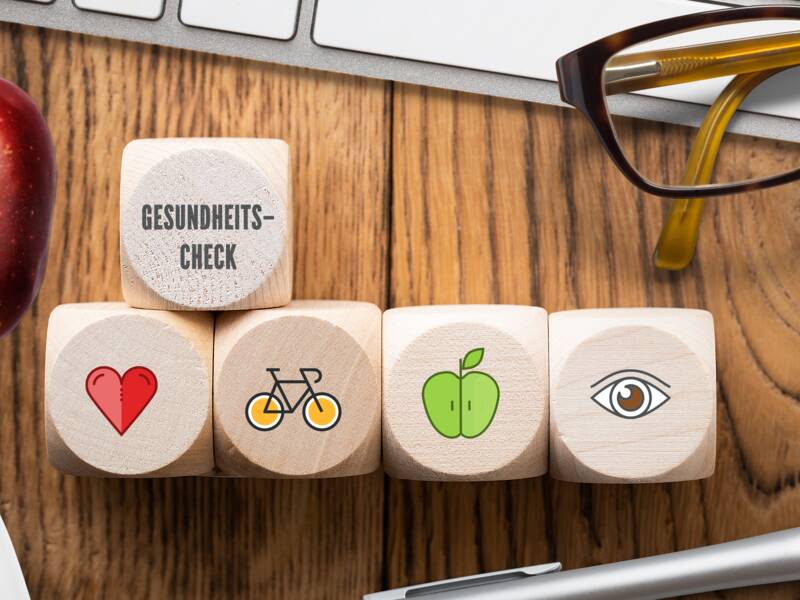 Fünf Würfel liegen auf einem Tisch mit einem Herz, Fahrrad, grünen Apfel, Auge und dem Text "Gesundheits Check" darauf. Außerdem liegen noch eine Tastatur, ein Apfel und eine Brille auf dem Tisch